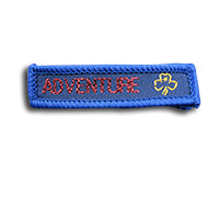 blue rectangle woven badge with the word adventure sewn in red in capital letters and a gold trefoil after the writing