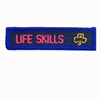 blue rectangle woven badge with the words life skills sewn in pink in capital letters and a gold trefoil after the writing