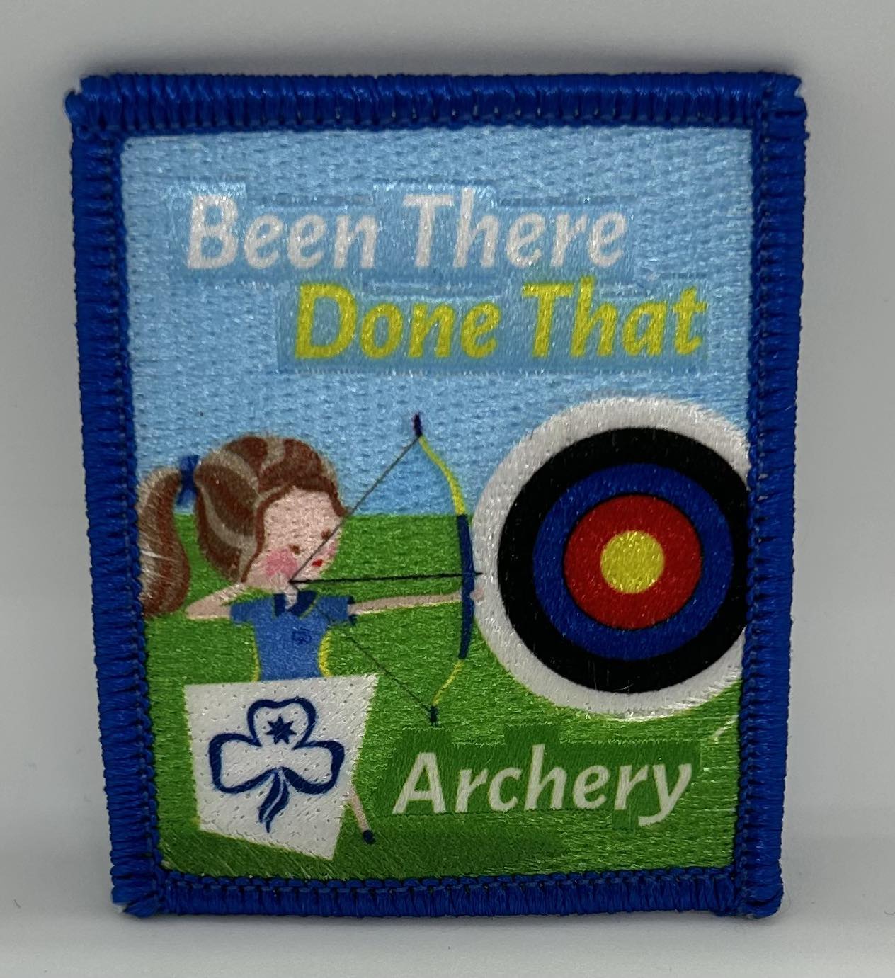 a square badge that is bound in blue with a guide in uniform pointing a bow and arrow  at a target