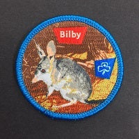 round badge bound with blue trim with a grey, brown and white Bilby on a brown, orange and yellow background