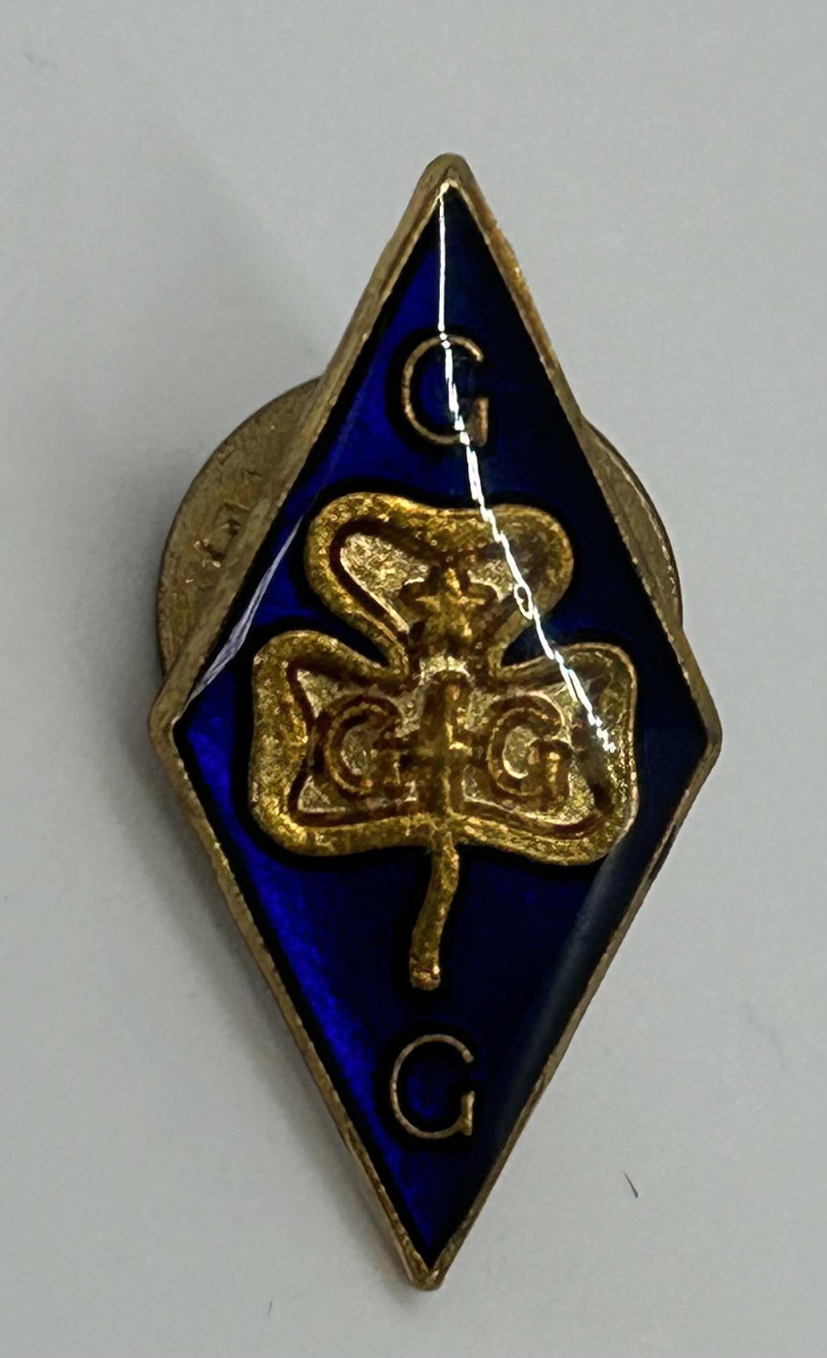 a diamond shaped blue enamel badge with a gold trefoil and the letters G G with a pin for pushing through a tie to secure