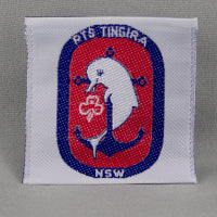 Square badge with white background. Badge features a dark blue oval with the words RTS Tingira NSW. Inside the oval is red with the RTS Tingira symbol of a white dolphin resting on a blue anchor