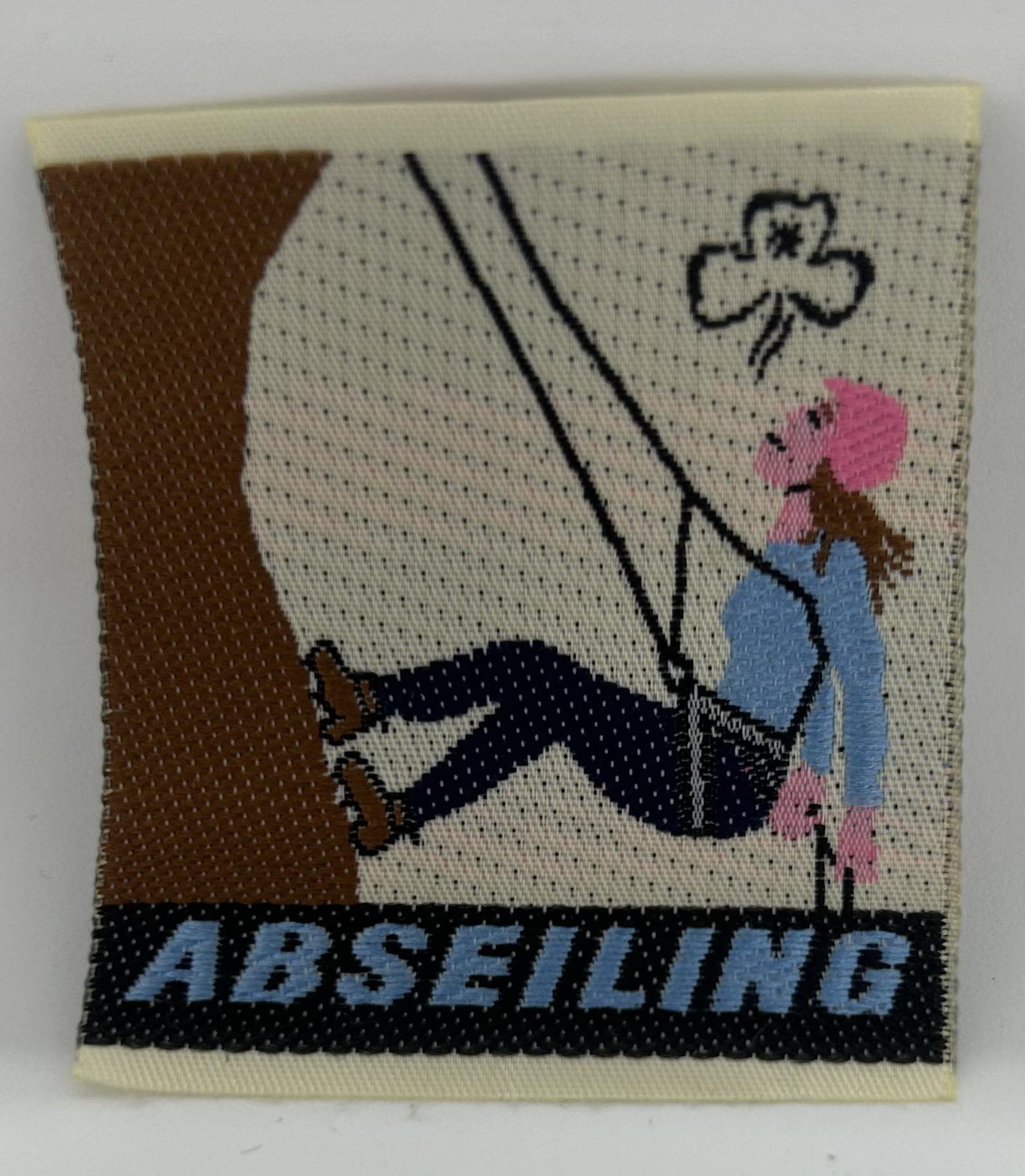 an unbound woven badge with a girl in a pink helmet abseiling down a rock face