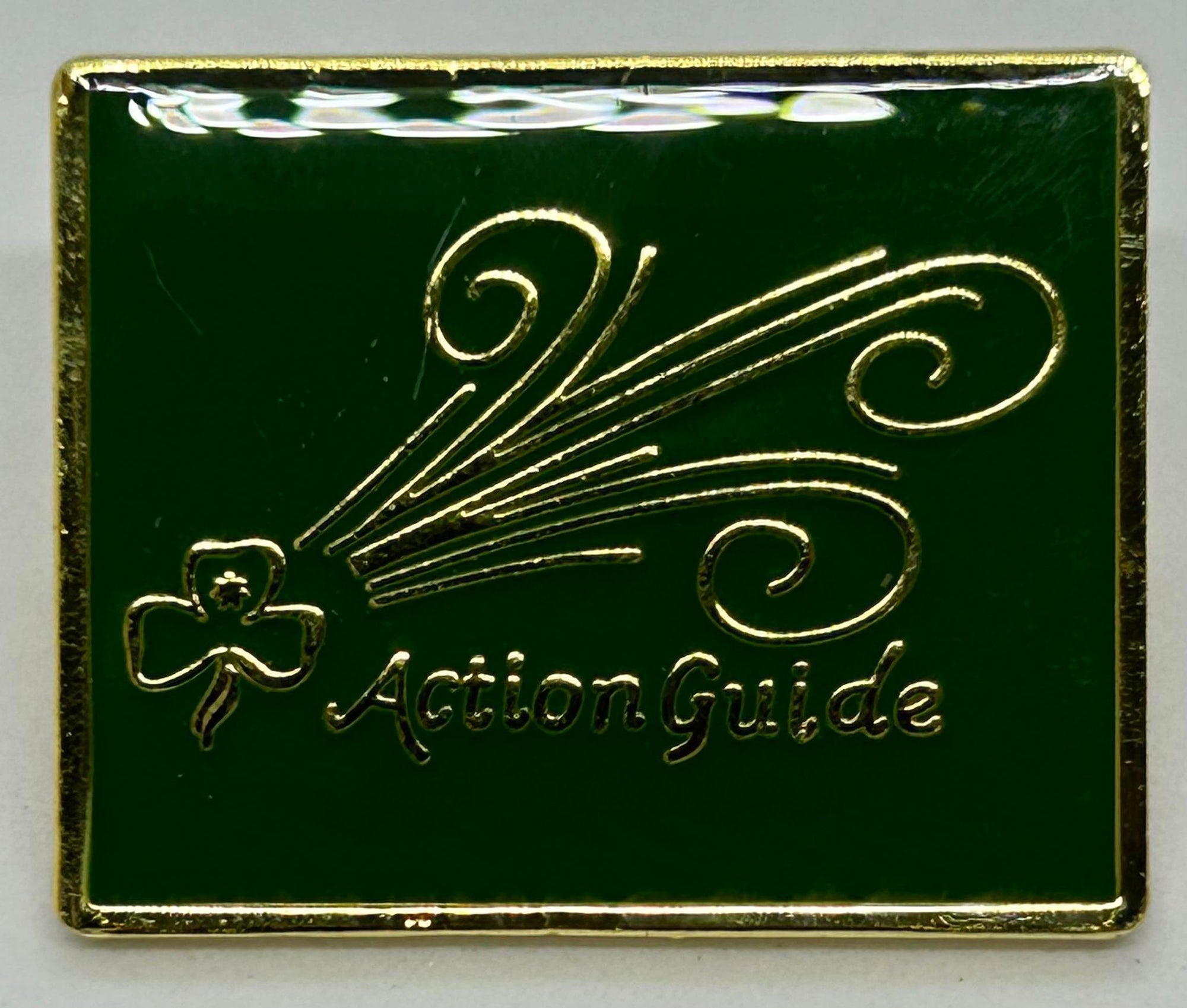 a green enamel badge with a trefoil, swirls and action guide in gold on the front