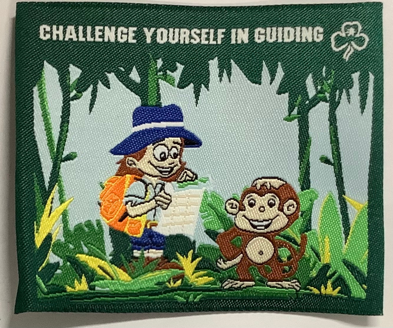 a square unbound badge with jungle trees, a monkey and a guide with a backpack