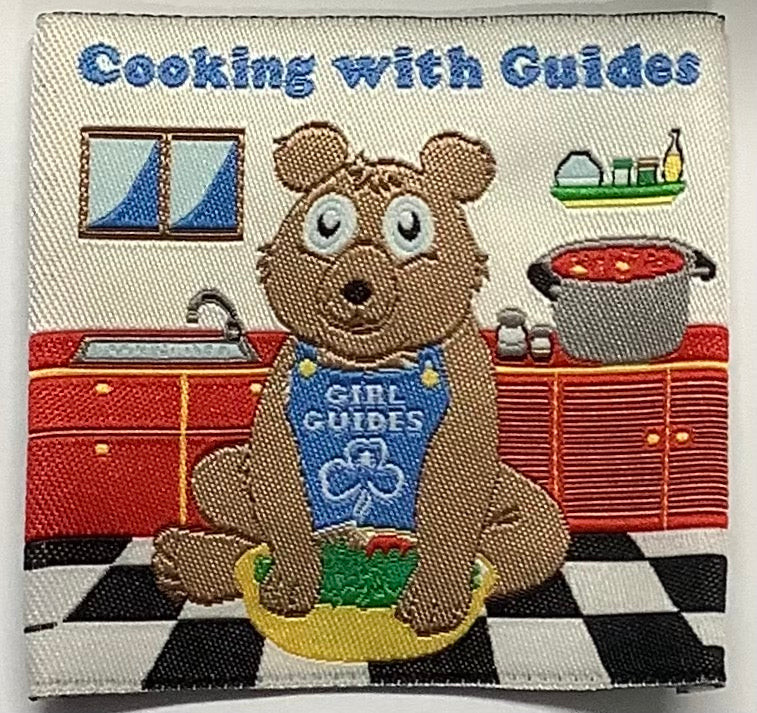 a square unbound badge with a kitchen scene and a bear with a girl guide apron on cooking