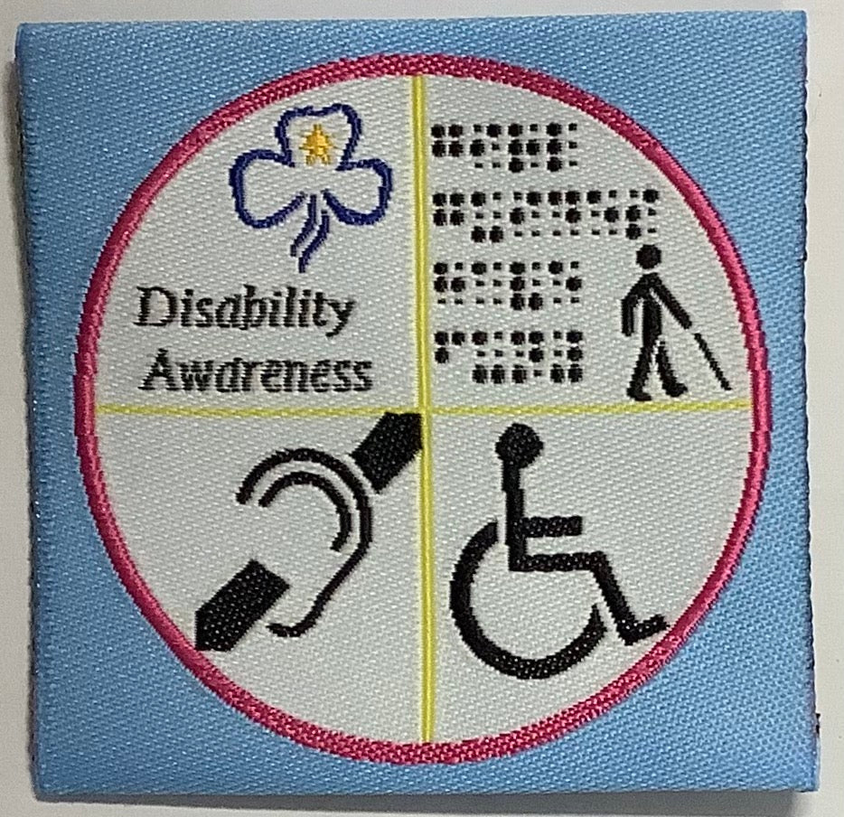 an unbound cloth badge with a light blue background with a white circle with a picture of a person with a cane, some braille, a hearing assist symbol and a wheel chair symbol