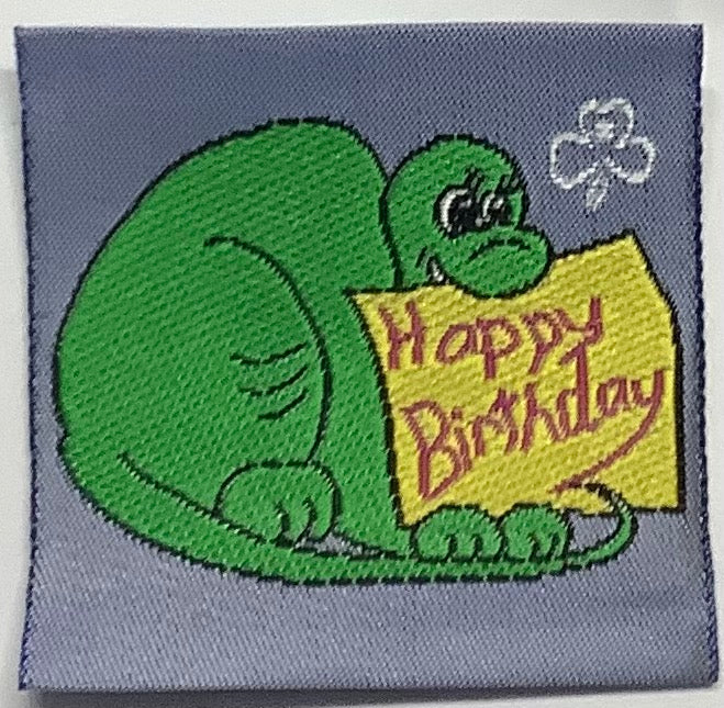 a square grey unbound badge with a green dinosaur with a yellow happy birthday sign in its mouth
