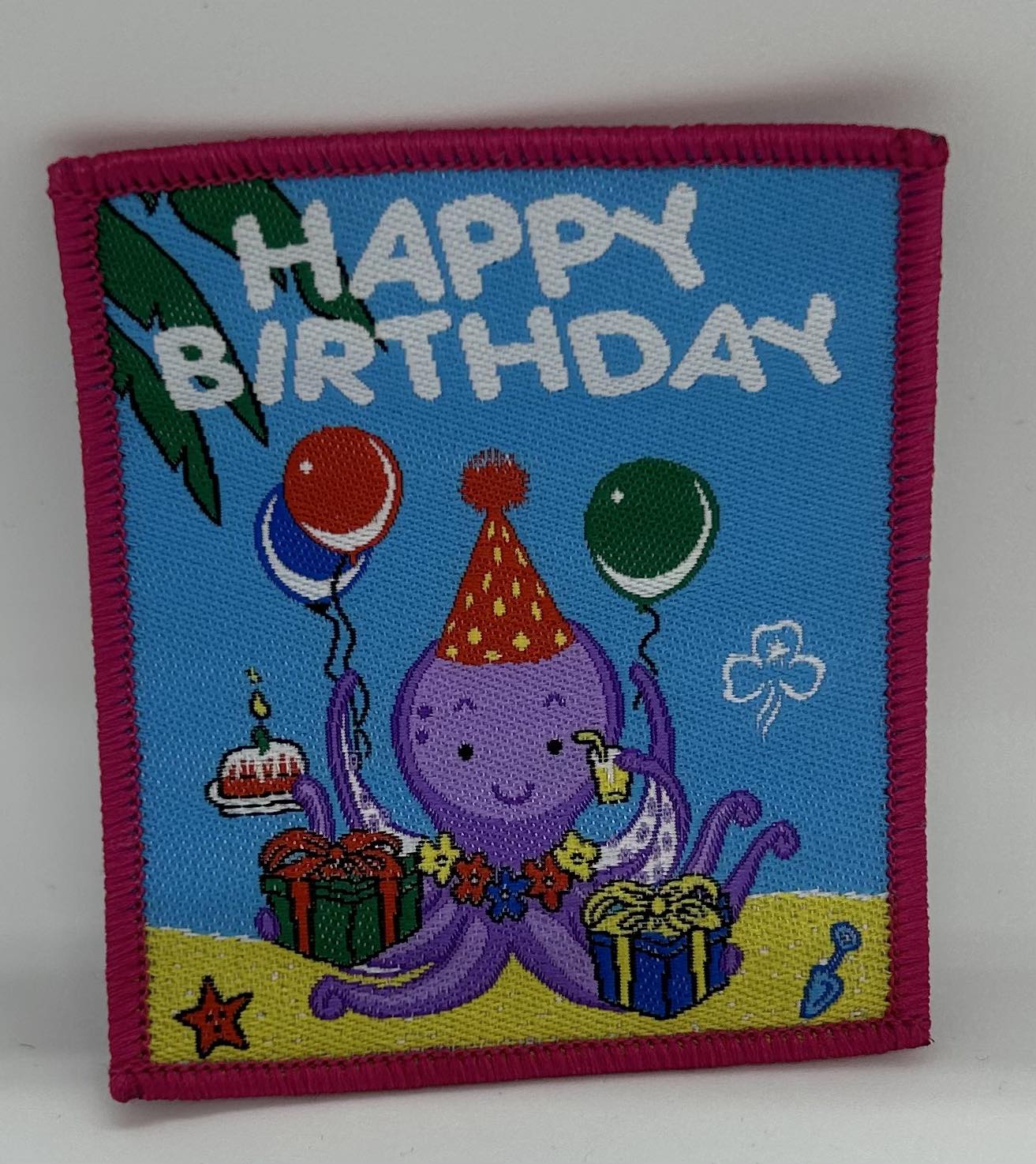 a square badge bound in red with an octopus with a party hat, balloons and presents