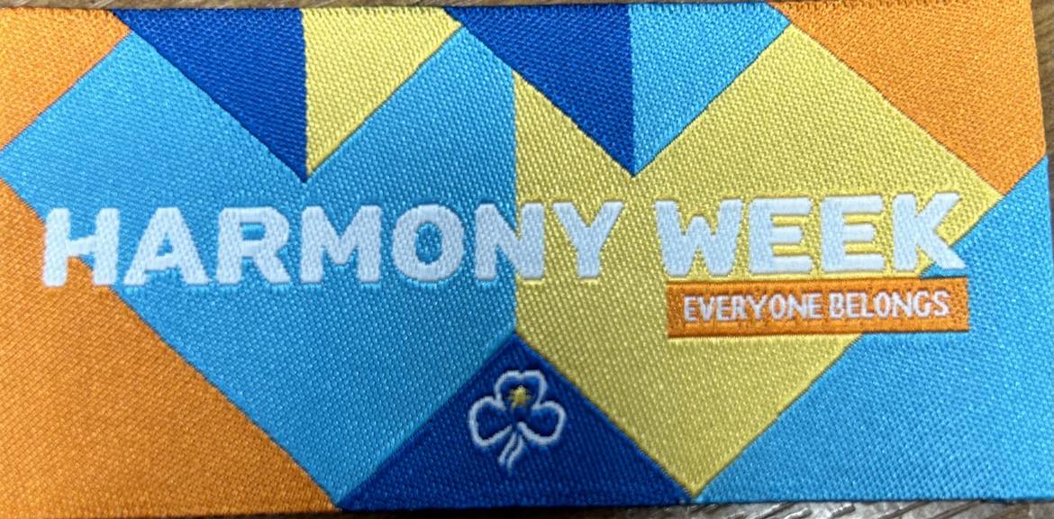 a rectangular unbound badge in blue, yellow and orange with angular shaped coloured patches with harmony week written in white in the middle of the badge