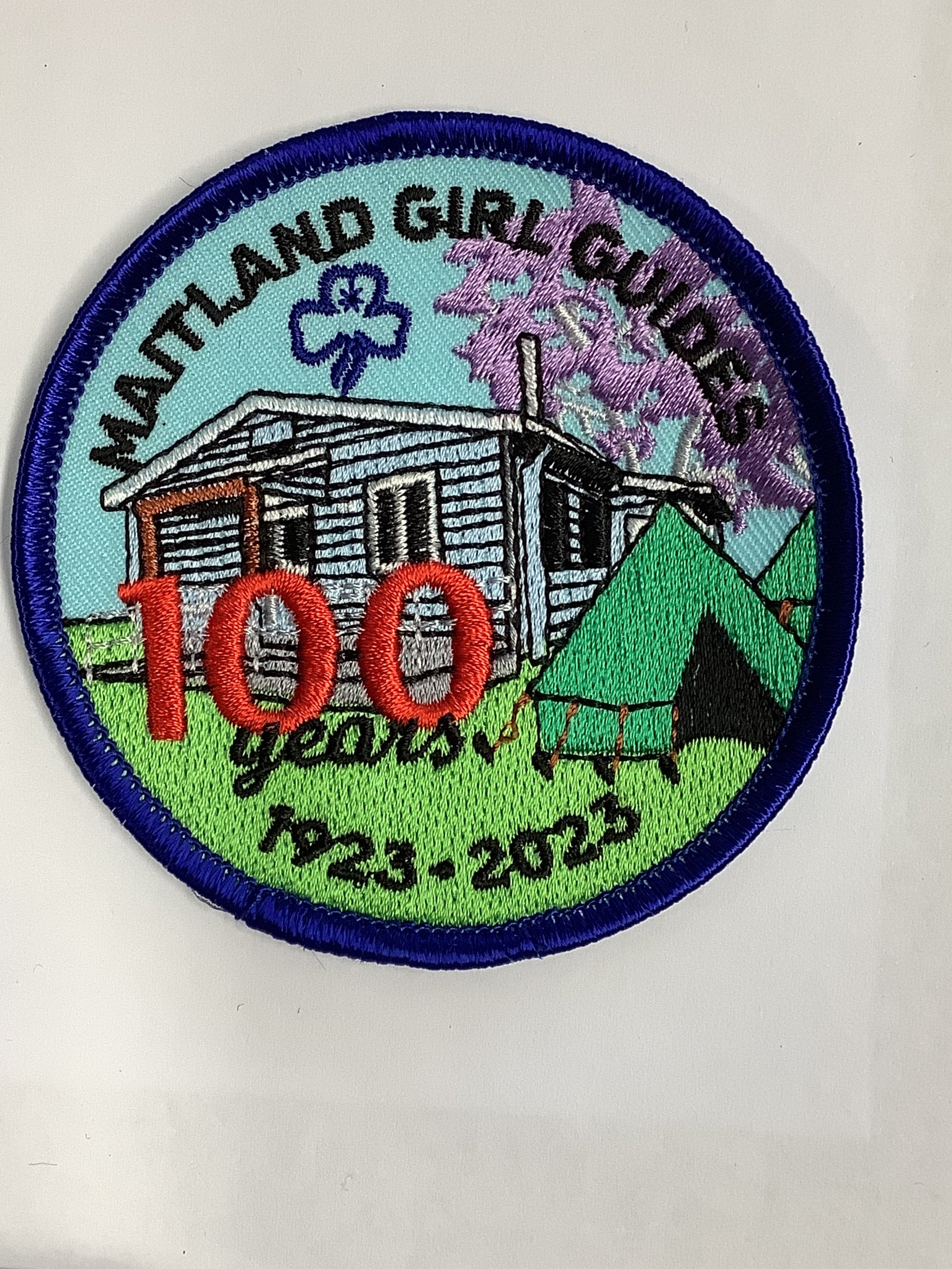 a round badge bound in blue with a tent and a picture of Maitland guide hall