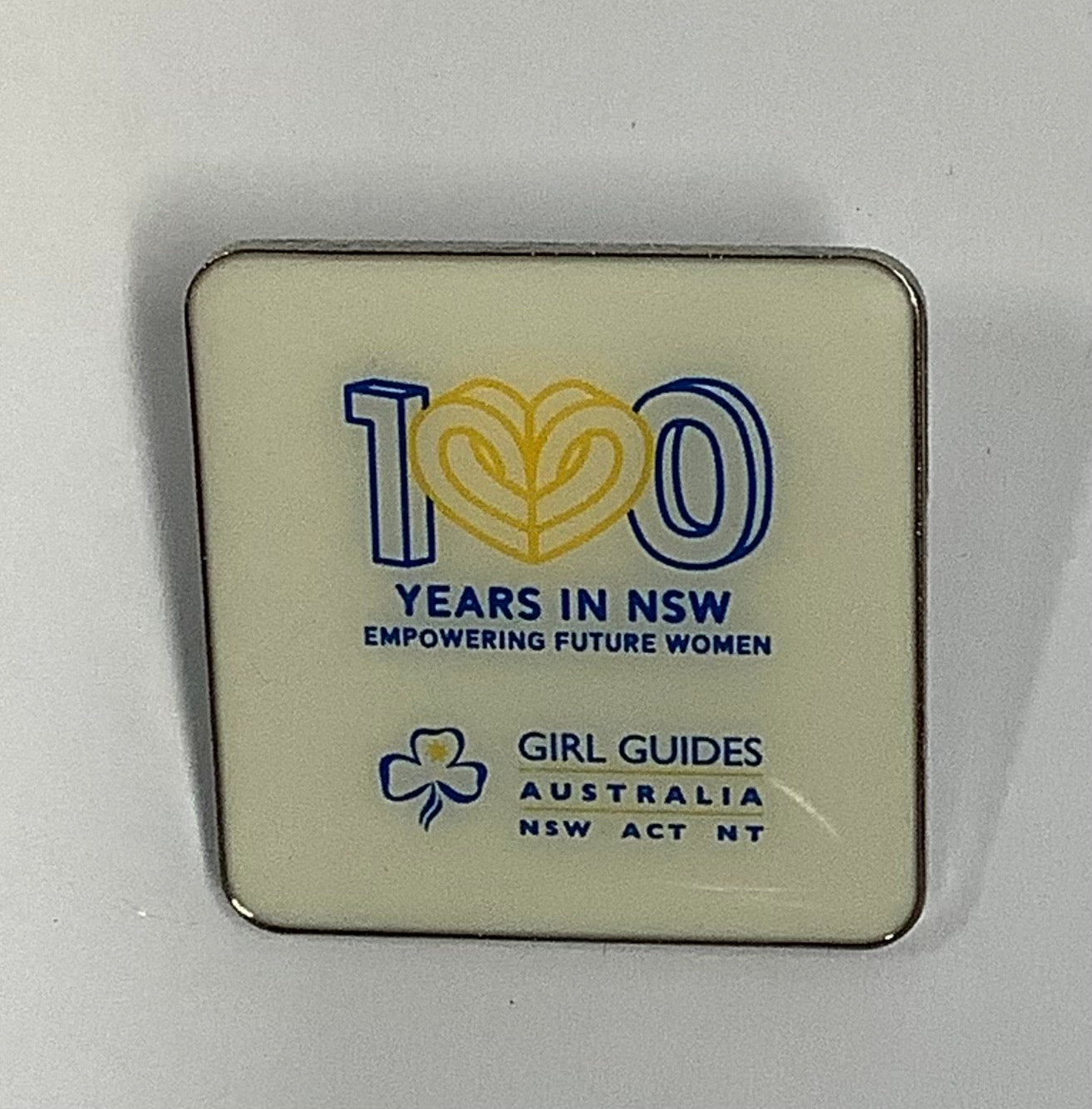 a square white enamel badge with the 100 years logo and the girl guides NSW, ACT & NT logo 