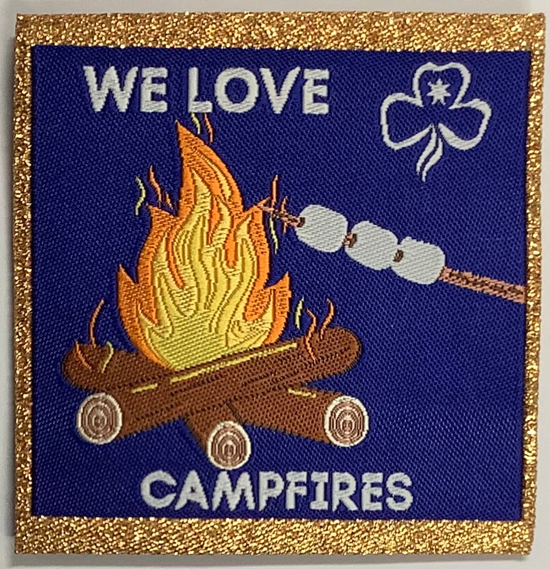 a square unbound badge with a campfire with marshmallows roasting on a stick with gold edging