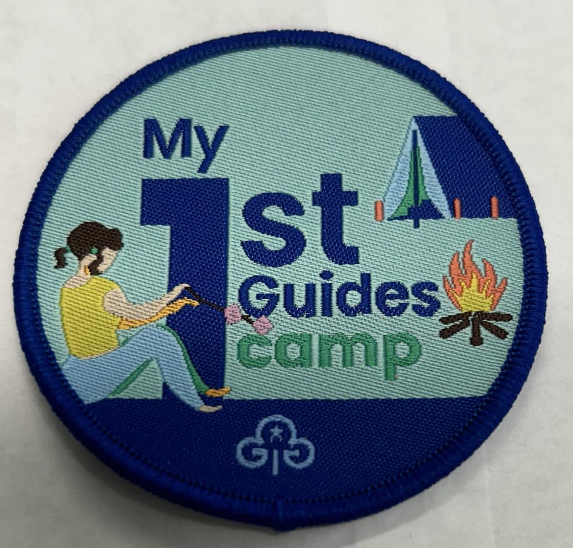 a round badge bound in dark blue with a girl toasting marshmallows, a campfire and a tent on it with the words my 1st guides camo