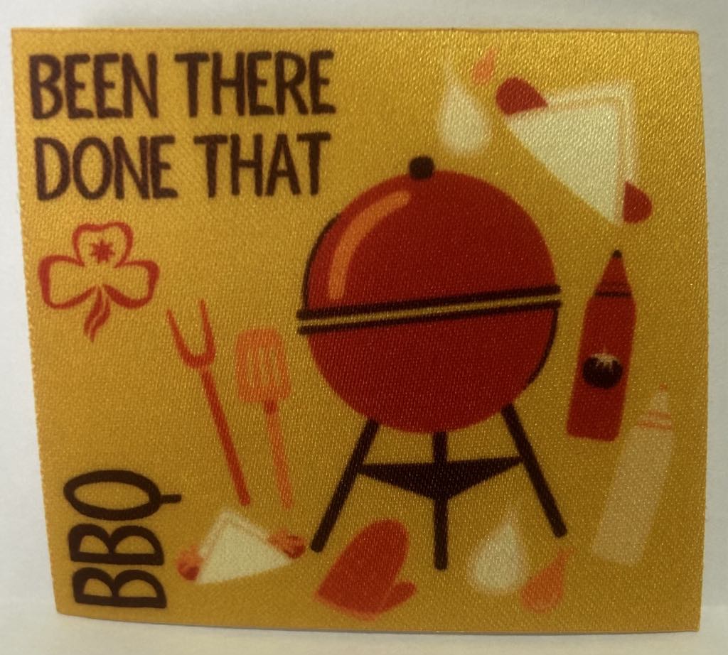 a square unbound badge that has a yellow background with a red bbq and utensils and sausage sandwich with the words been there done that BBQ in brown