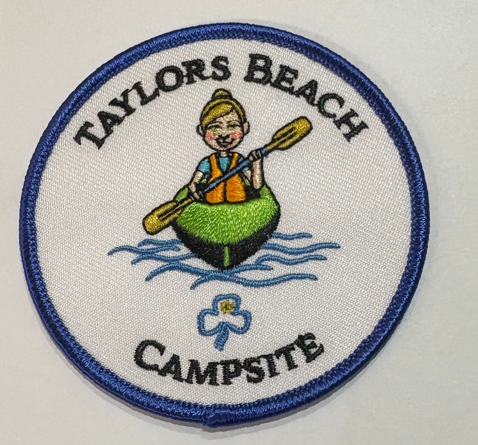 A round white badge bound in blue with the words Taylors Beach Campsite and a girl in a canoe