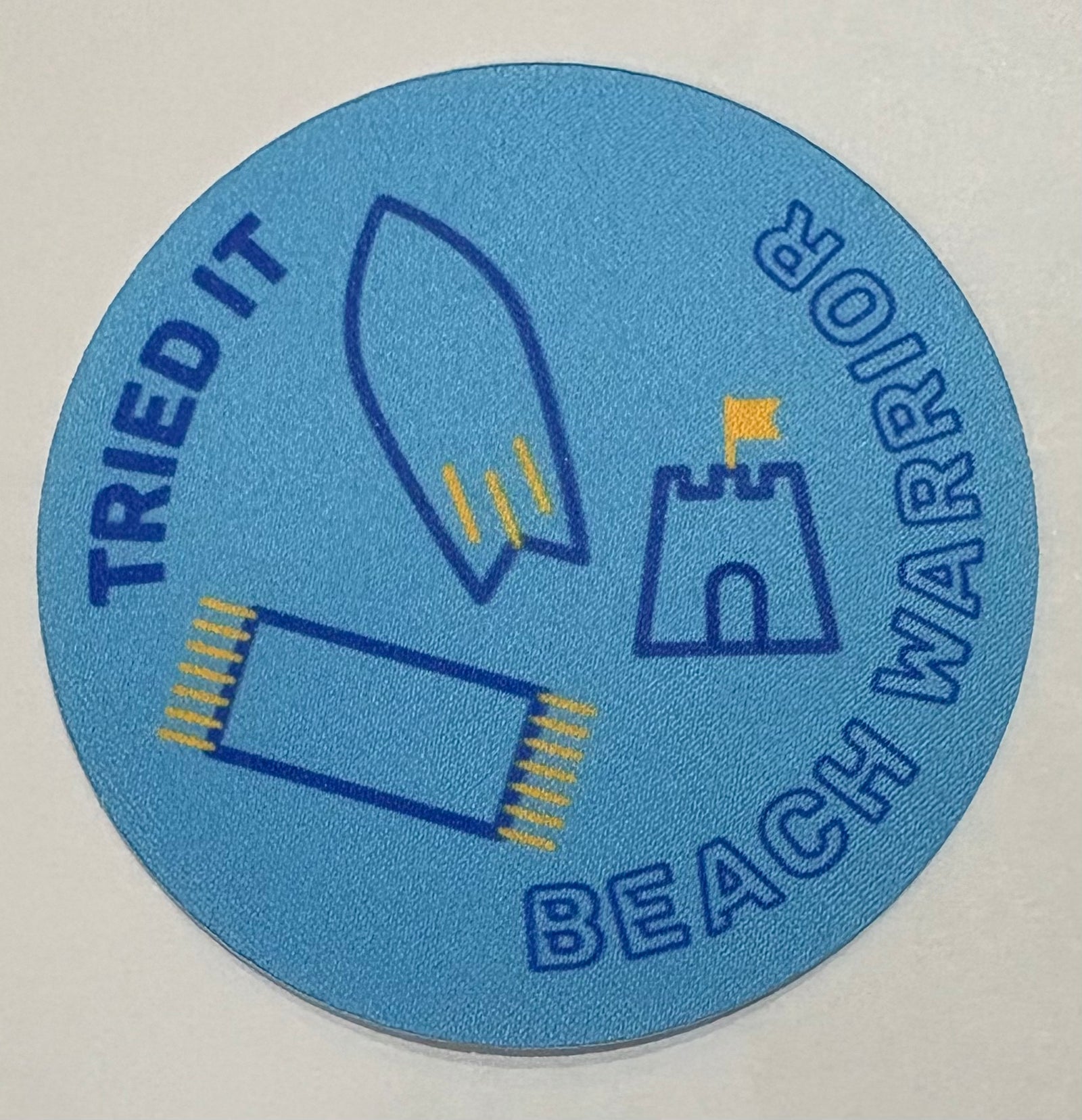 A round blue unbound badge with a towel, surfboard and a sand castle 