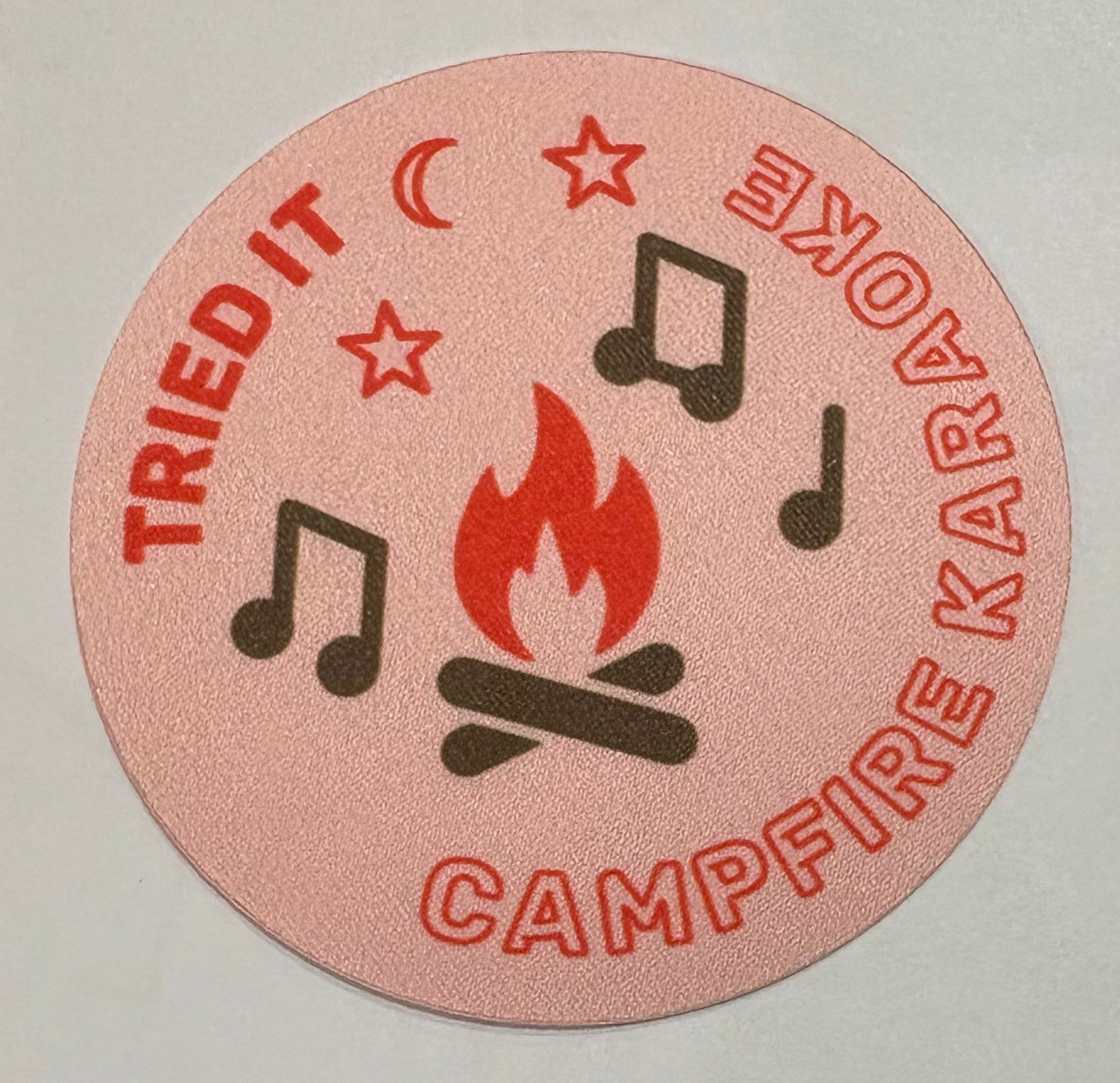 A round light pink unbound badge with a campfire and some musical notes