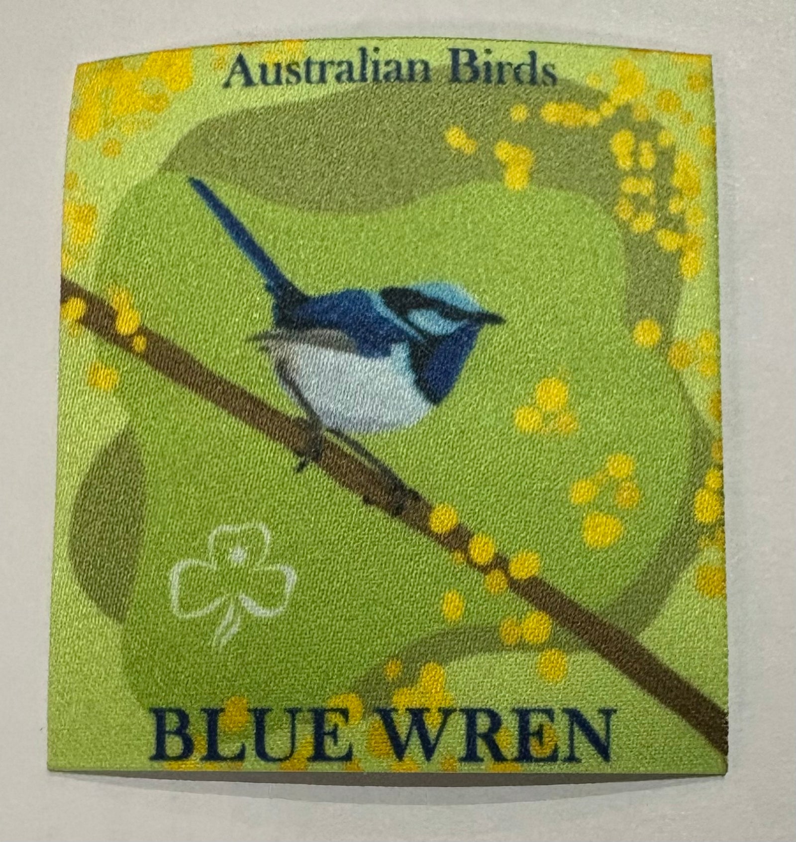 A square green unbound badge with a blue wren sitting on a branch with some wattle in the background 