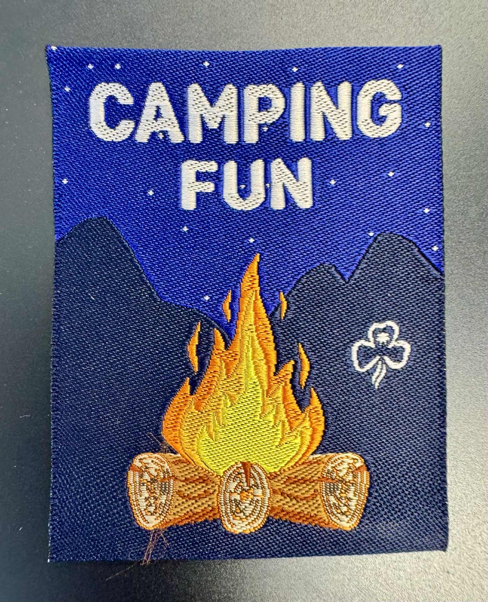 A rectangular unbound badge with a campfire on a dark blue background 