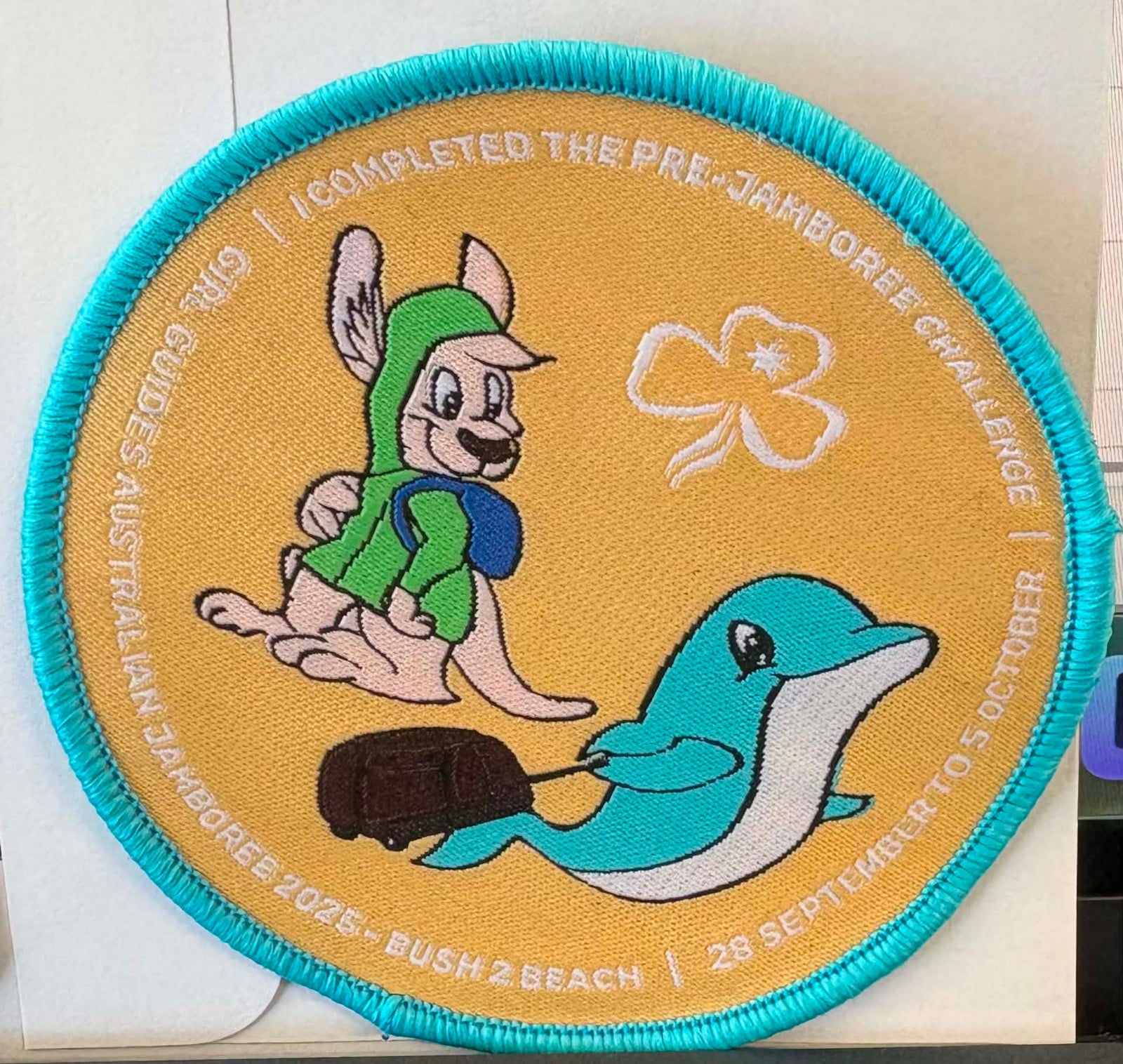 A circular embroidered badge with a turquoise border on a tan background, featuring a kangaroo in a green shirt and blue backpack, a white clover, a blue-and-white dolphin, and white text around the edge reading 'I COMPLETED THE PRE-JAMBOREE CHALLENGE' and 'GUIDES AUSTRALIA 2025 - BUSH 2 BEACH | 28 SEPTEMBER TO 5 OCTOBER'.