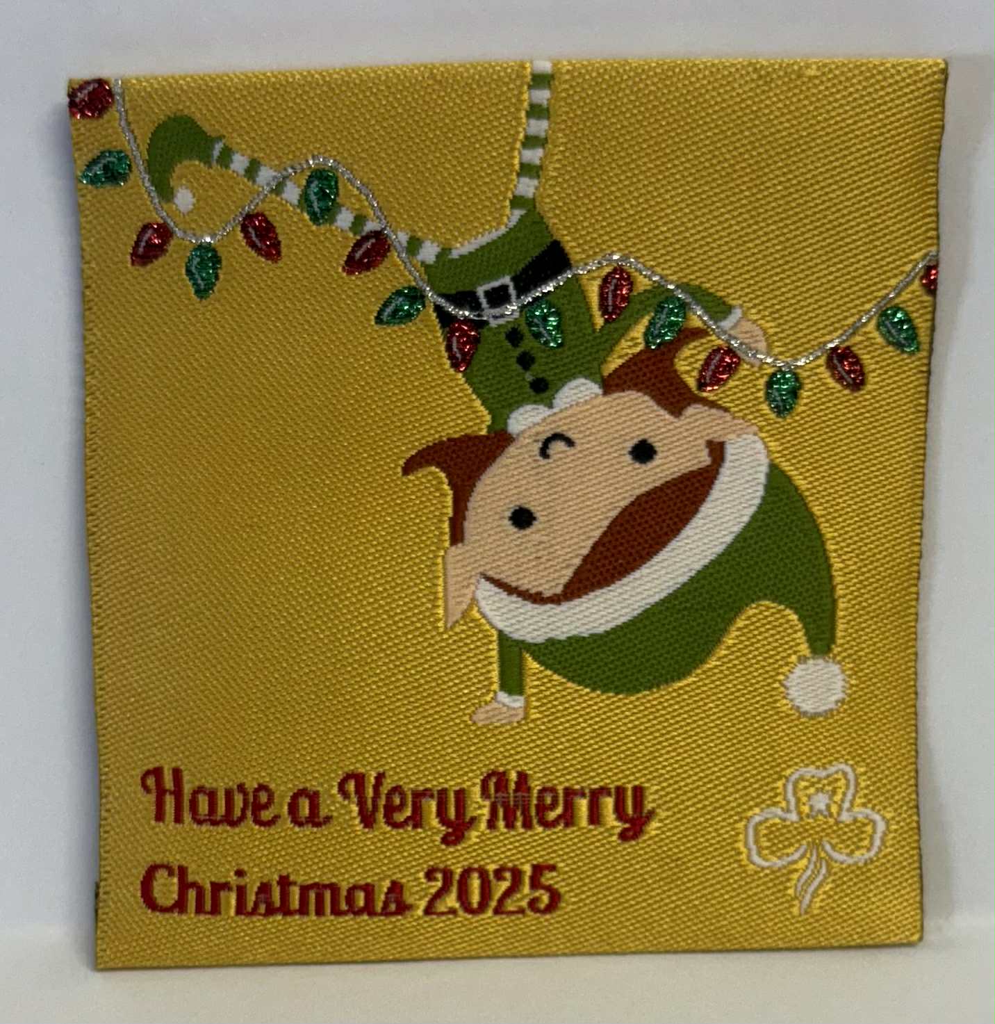 Decorative badge with a doll in an elf outfit and Christmas lights on a yellow background.