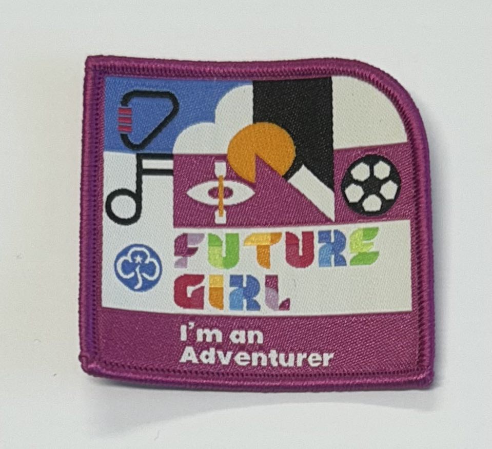 a square badge bound in magenta with fire girl I'm an adventurer written on it with some sports equipment and a musical note