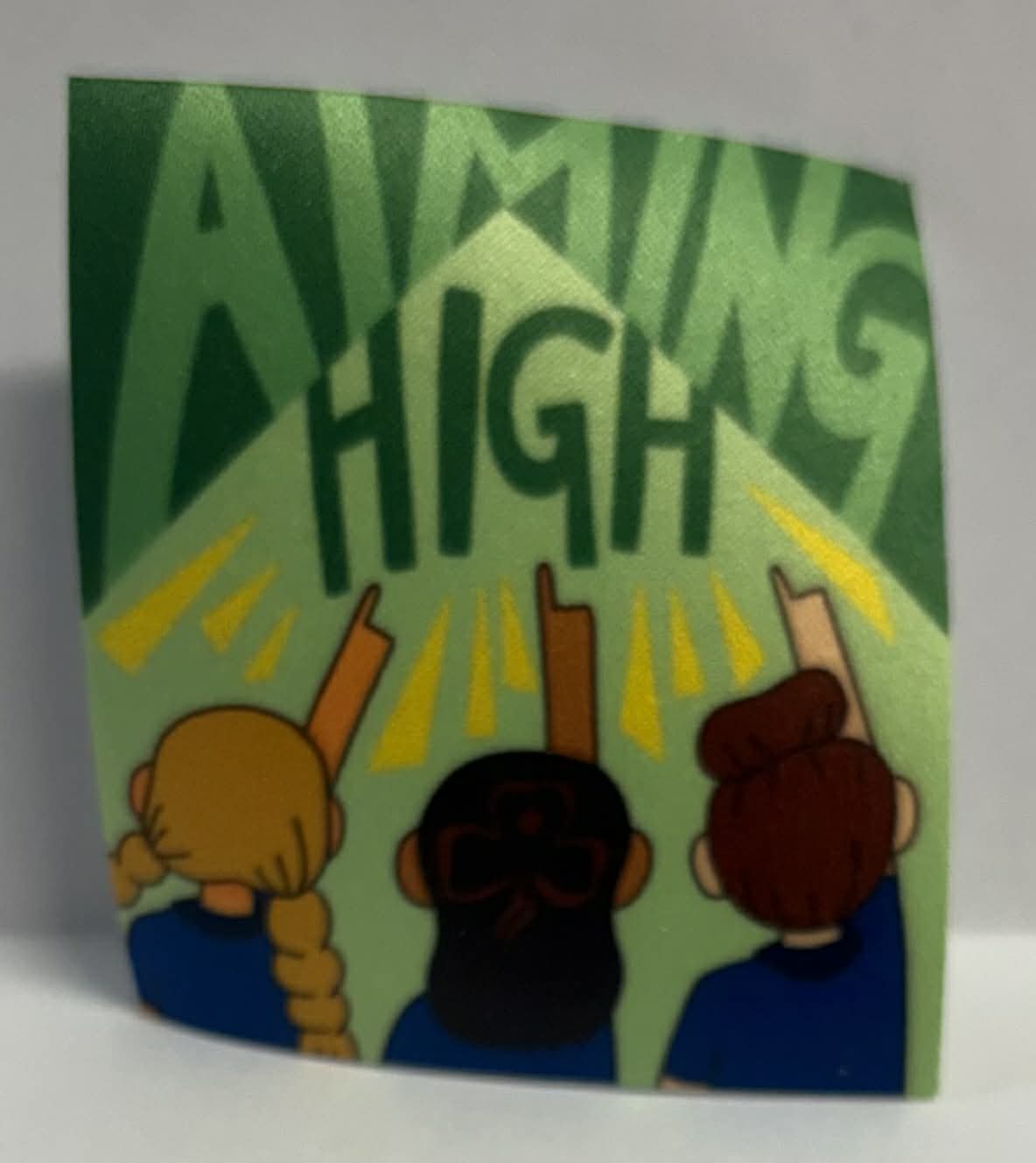 Square unbound badge with three people pointing upwards and 'Aiming High' text on a green background