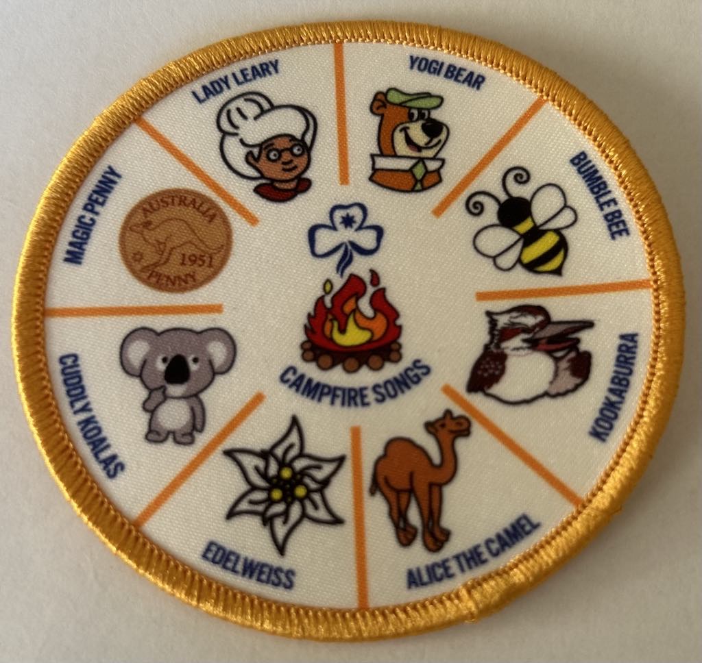 a round white badge bound in gold with a campfire in the middle with 8 campfire song titles around the outer edge