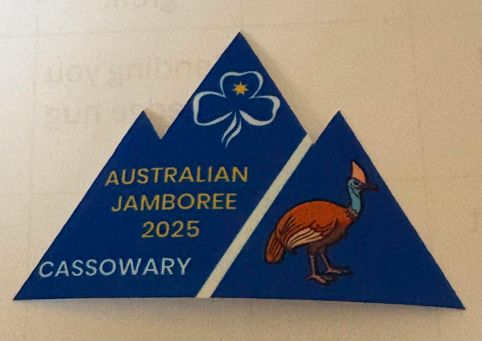 A triangular blue commemorative badge for the Australian Jamboree 2025 Cassowary, featuring gold and white text, a white trefoil with a yellow star, and a brown cassowary bird.