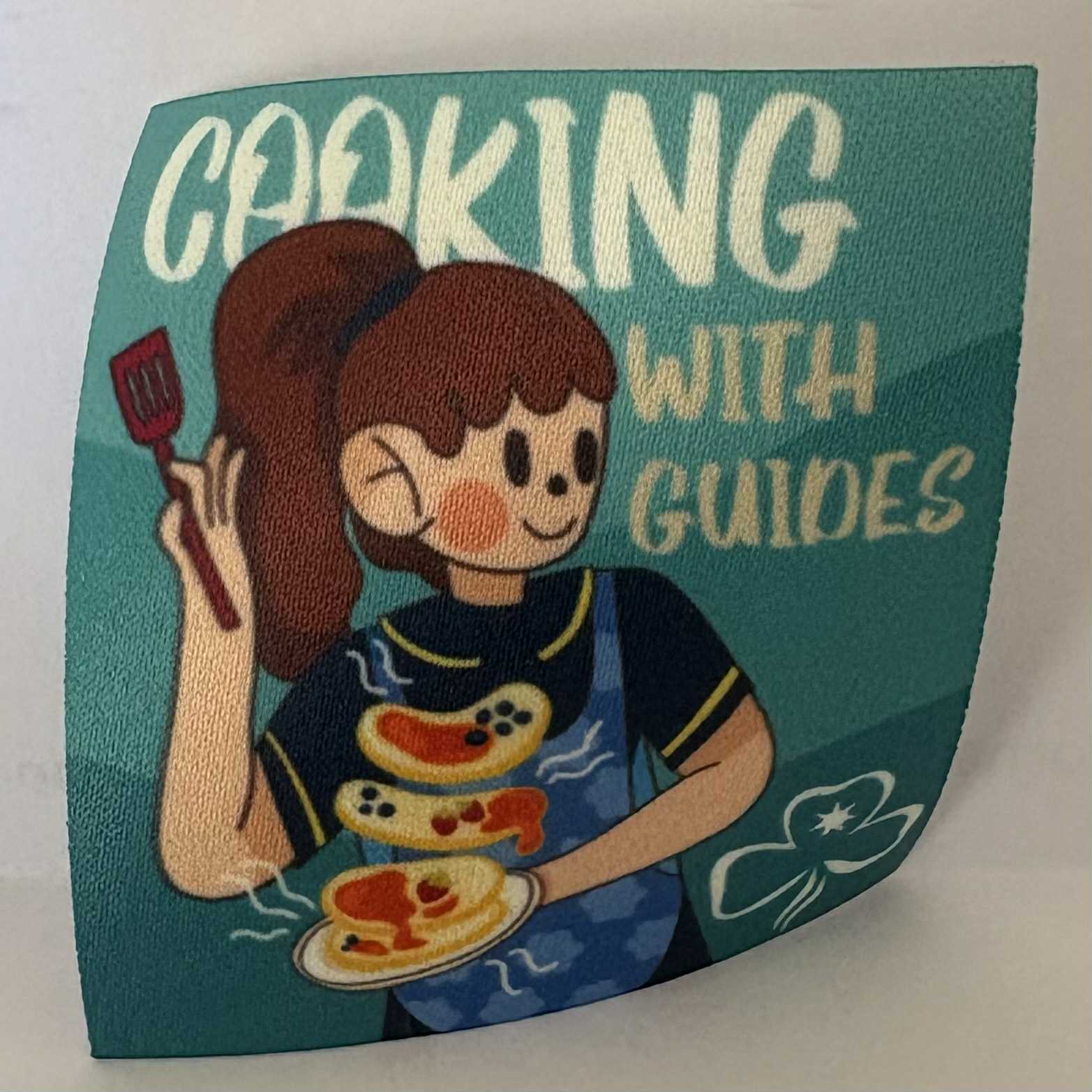 a square unbound badge with a girl holding a spatula and plate, and text 'Cooking with Guides' on a teal background.