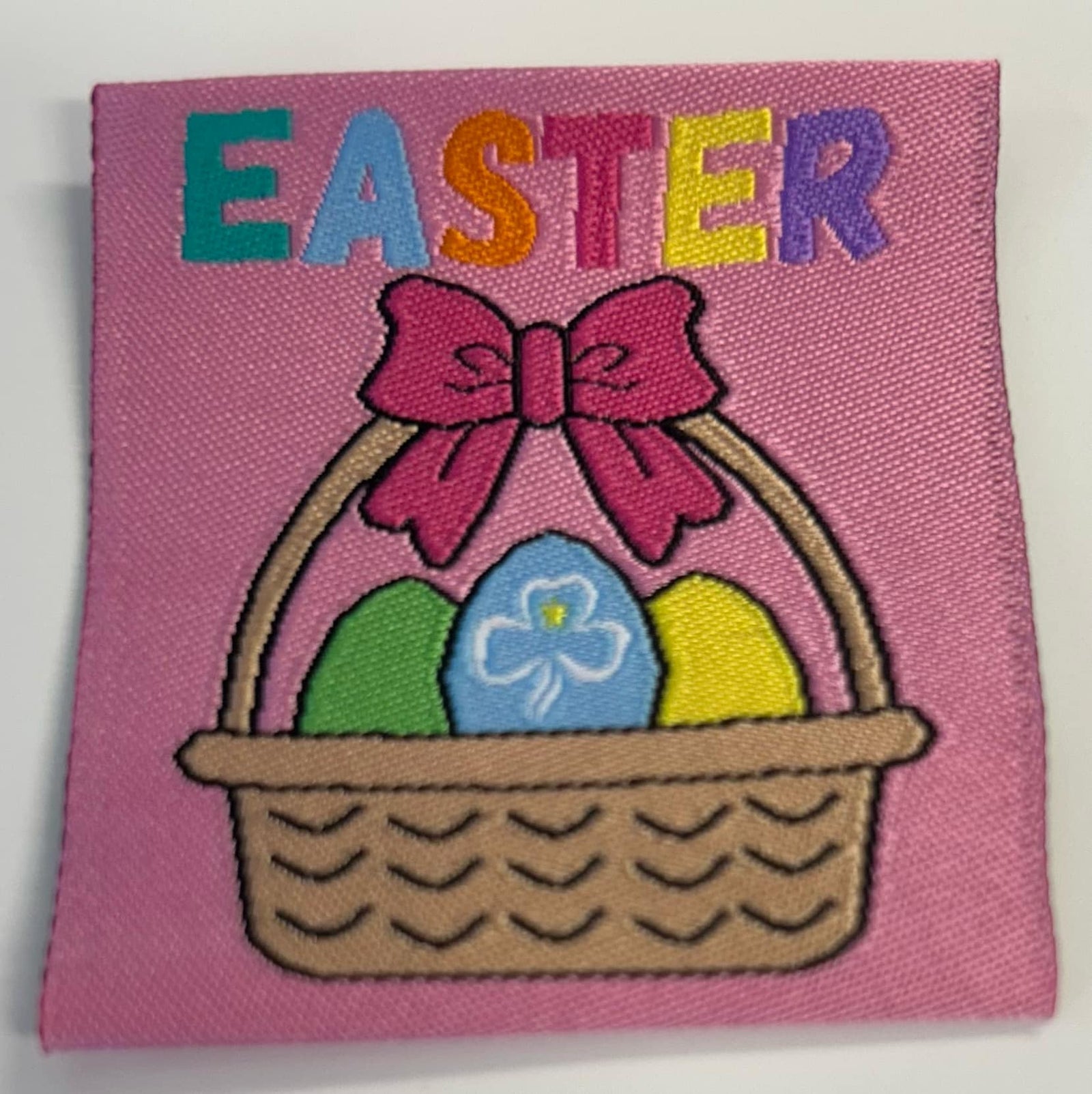 a pink square unbound badge with a basket with eggs in it and a pink bow