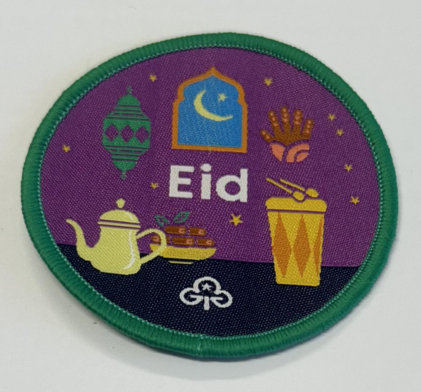 a round badge bound in green with a purple and dark blue background with drums, teapot, food, lantern and the moon with the word Eid