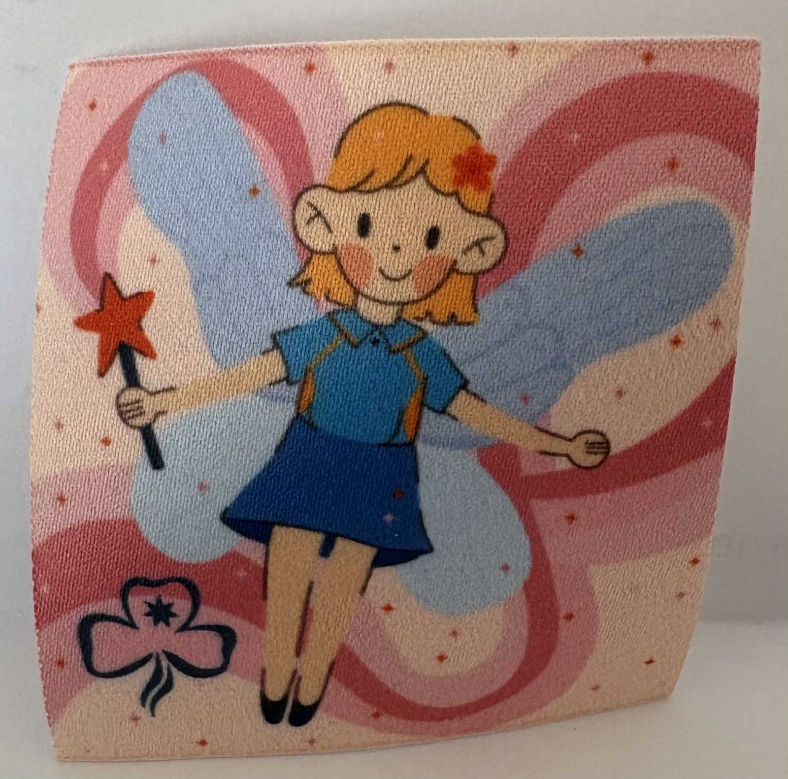a square unbound badge with a Fairy character with blue wings and a wand on a pink background