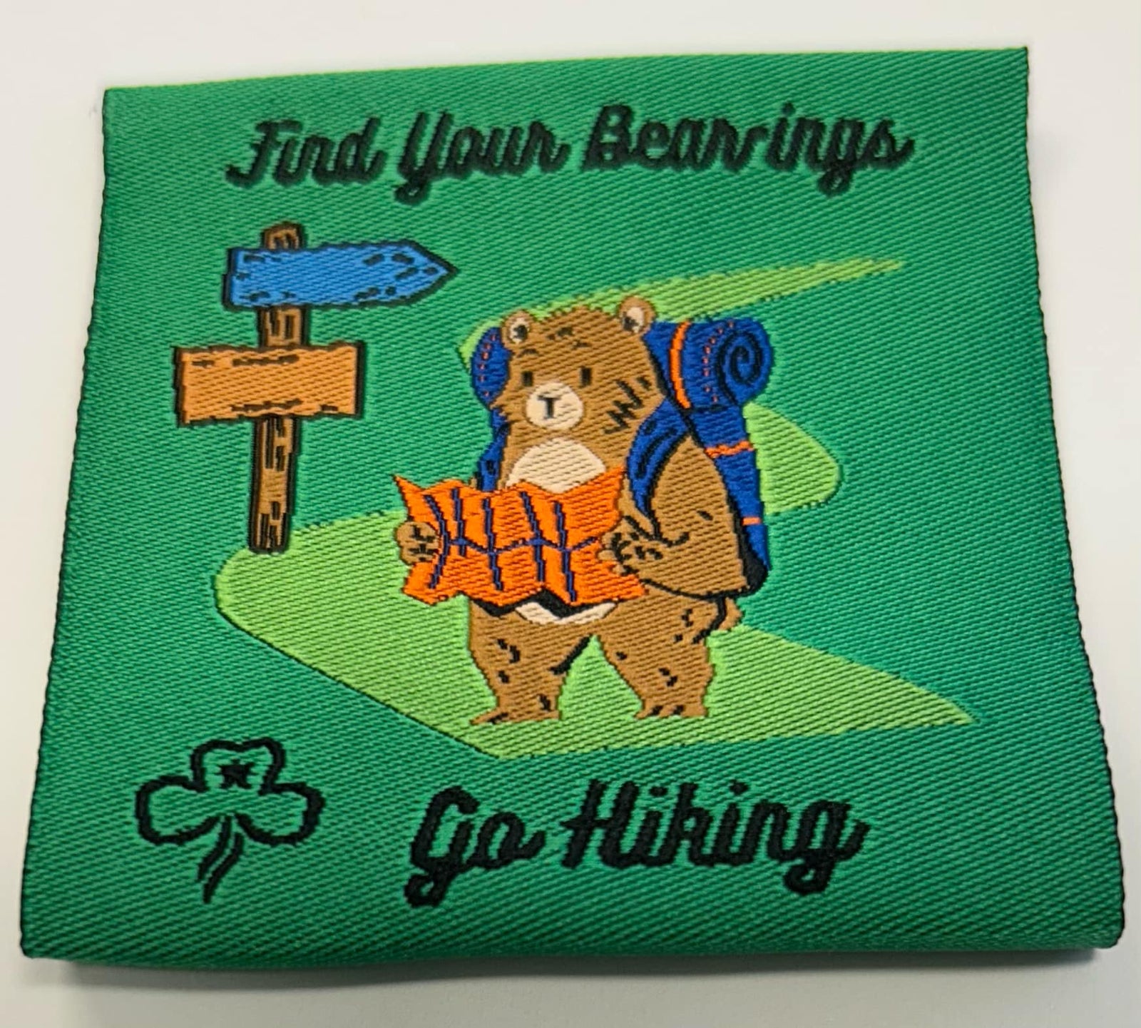 a square green unbound badge with a bear wearing a back pack holding a map near a sign post
