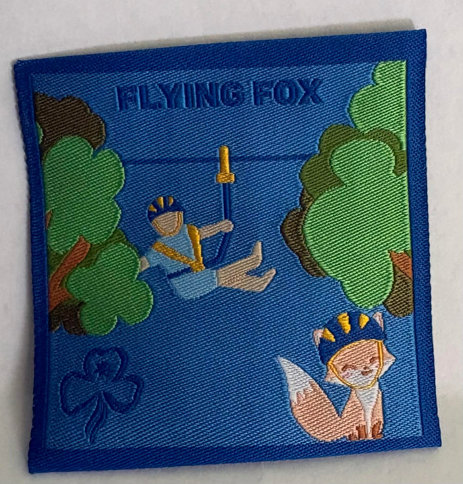 a square blue unbound badge with a girl on a flying fox in the trees and a fox cartoon character in a helmet on the ground