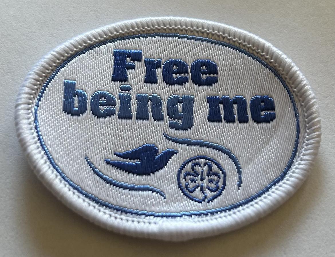 on oval shaped white badge that is bound in white and has free being me, a dove and the world trefoil in blue on the front