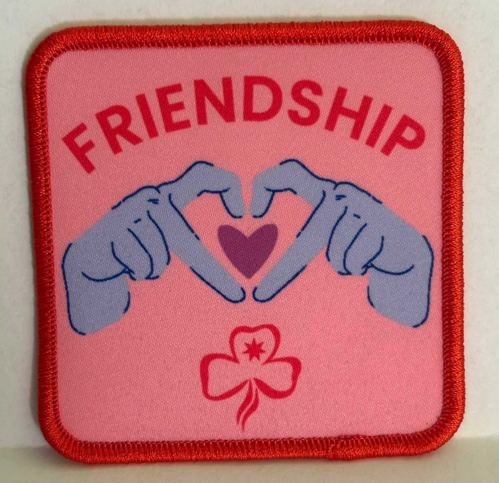 square pink bound badge with hands forming a heart and the word 'Friendship' 