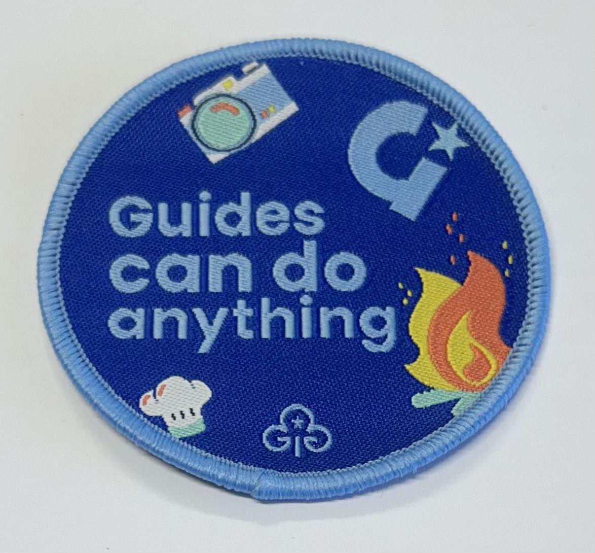 a round blue badge bound in light blue with the words guides can do anything on it with  a camera, campfire and chefs hat