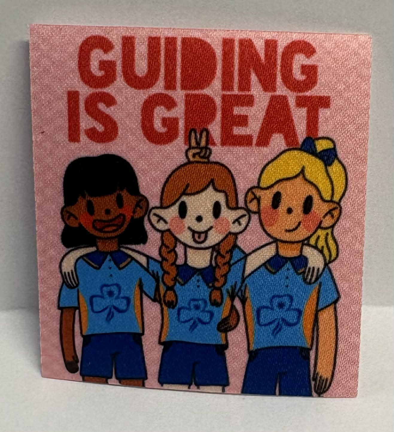 Square unbound badge with three cartoon girls in blue uniforms and 'Guiding is Great' text on a pink background.