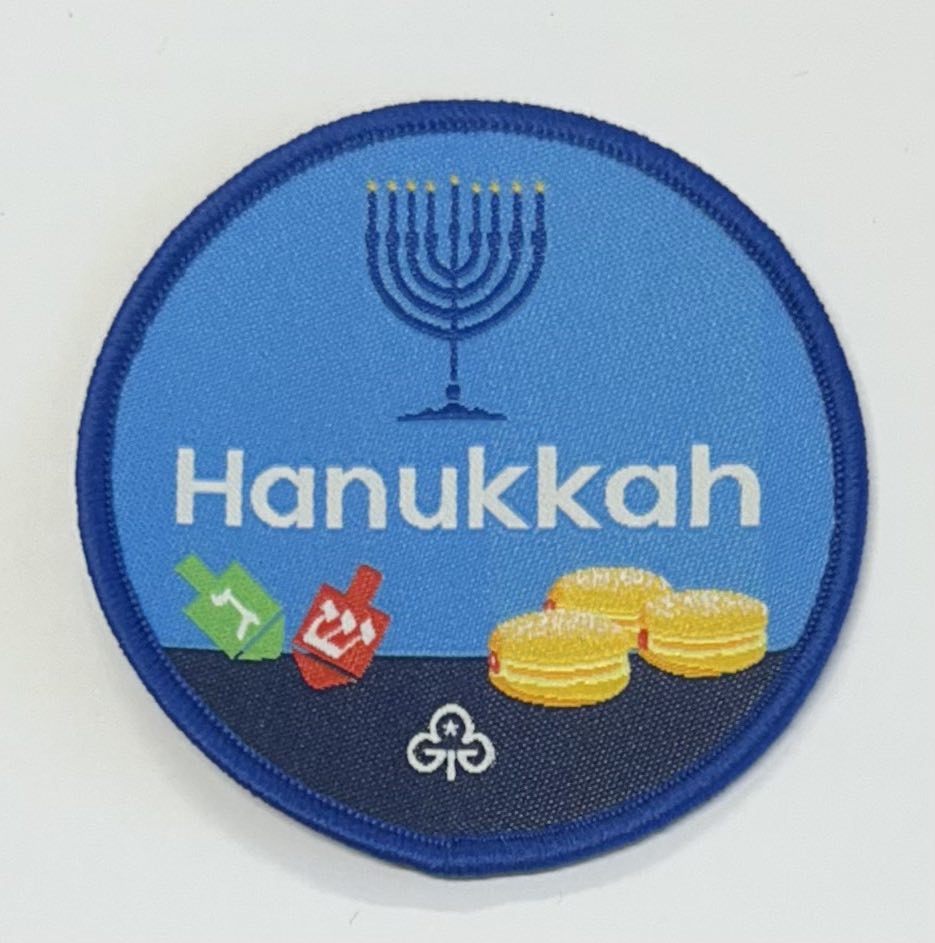 a round badge bound in blue with a two tone blue background with the nine branched candelabra and food on it with the word Hanukkah