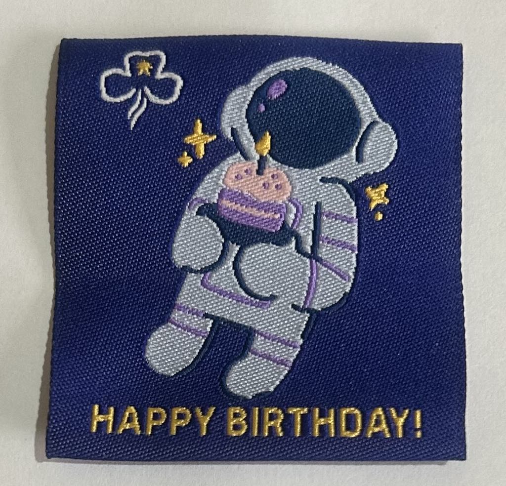a square unbound badge with a navy blue background with a astronaut holding a piece of cake with a candle in it