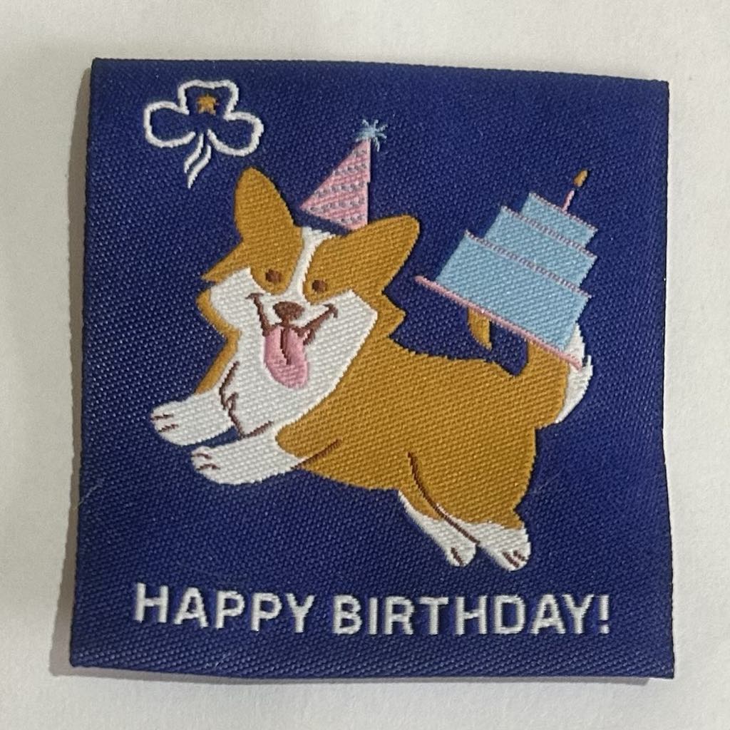 a square unbound badge with a blue background with a corgi with a party hat on and a cake on its tail and the words happy birthday