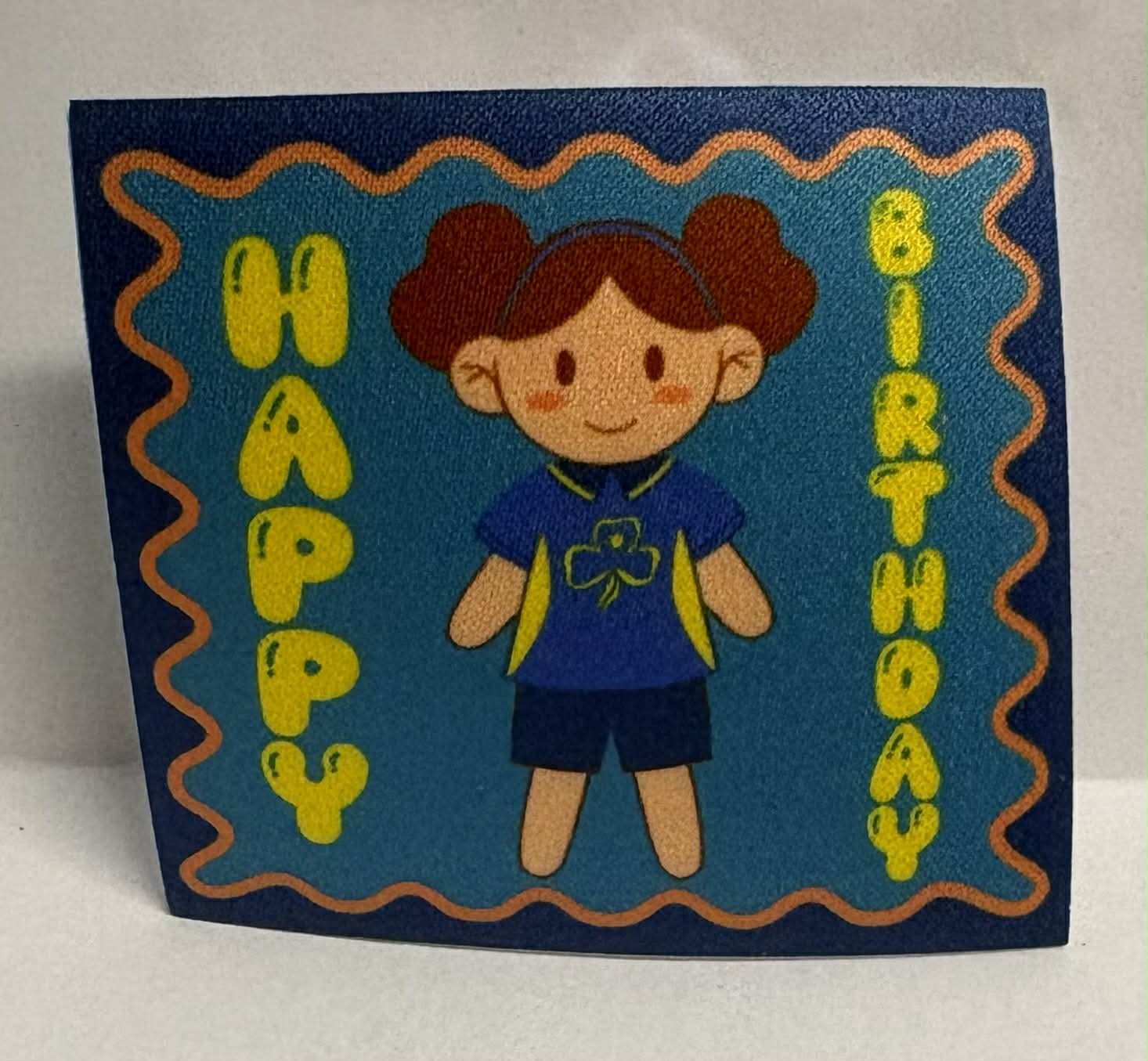 Square unbound badge with a cartoon character of a Girl Guide and 'Happy Birthday' text on a blue background