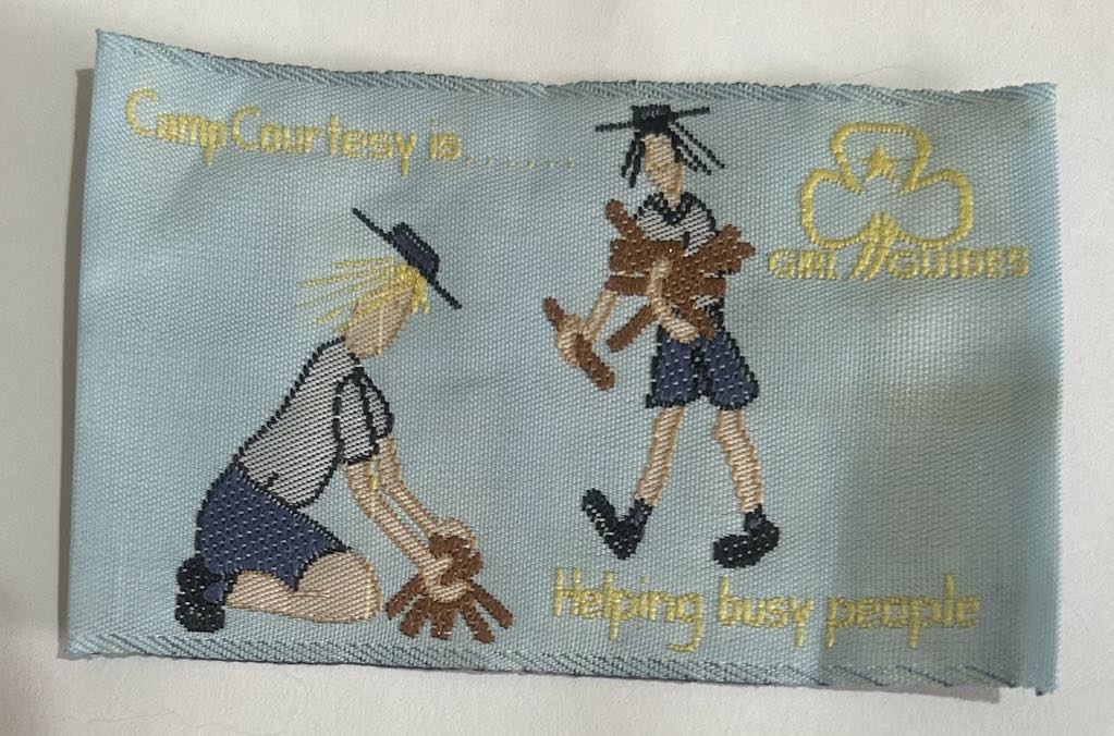 a rectangular unbound badge with a light blue background with two guides helping each other build a fire with the words camp courtesy is helping busy people