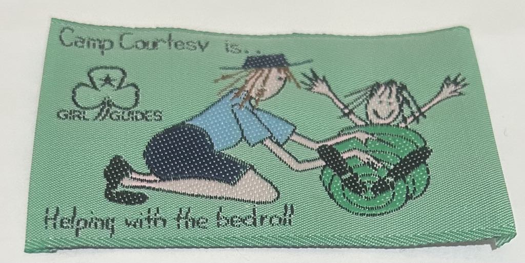 a rectangular unbound badge with a mint green background with one guide rolling another guide in a bedroll with the words camp courtesy is helping with the bedroll