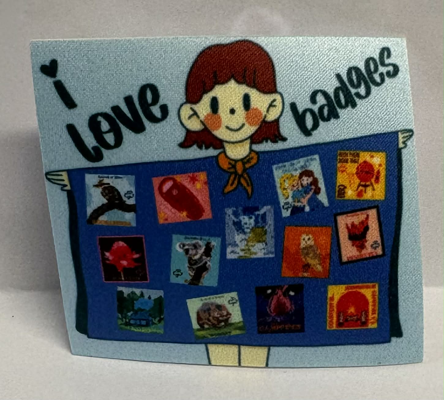 Square unbound badge with a cartoon character girl guide and 'I love badges' text, featuring various small images of our badges on a blanket on a blue background.