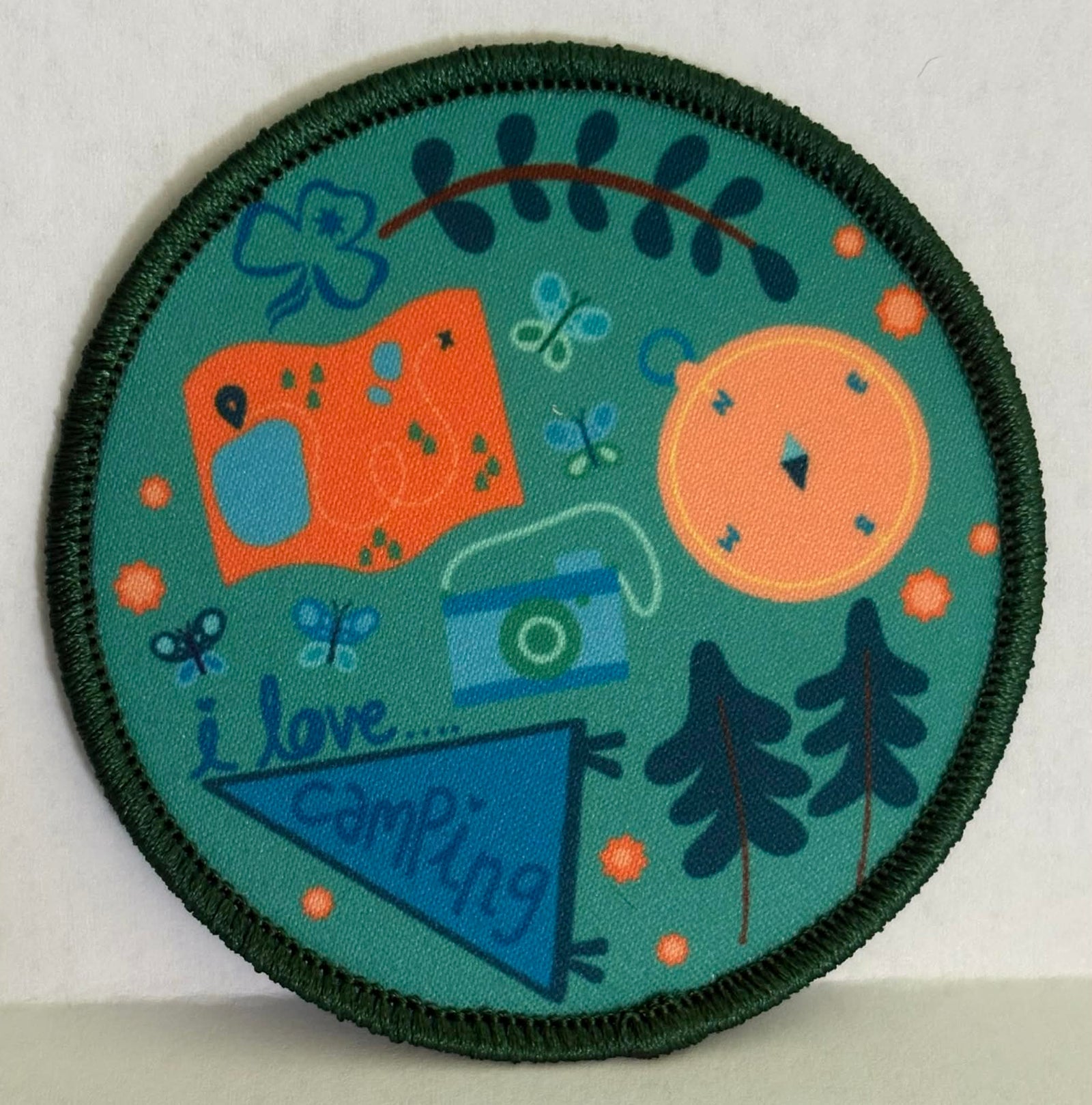 a round and bound badge with a compass, camera, trees, map and tent on a green background