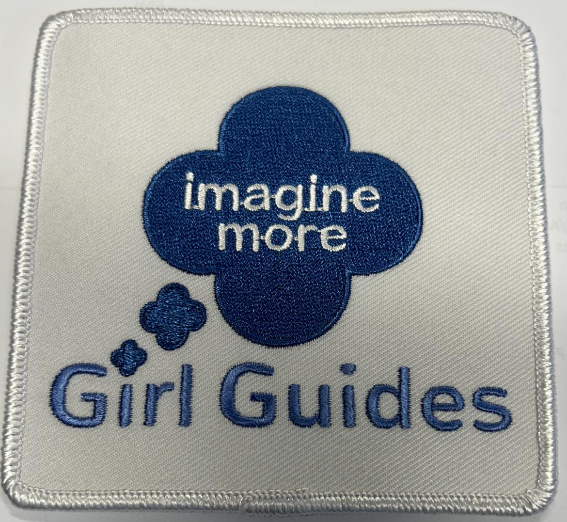 a square badge bound in white with the words imagine more girl guides on the front