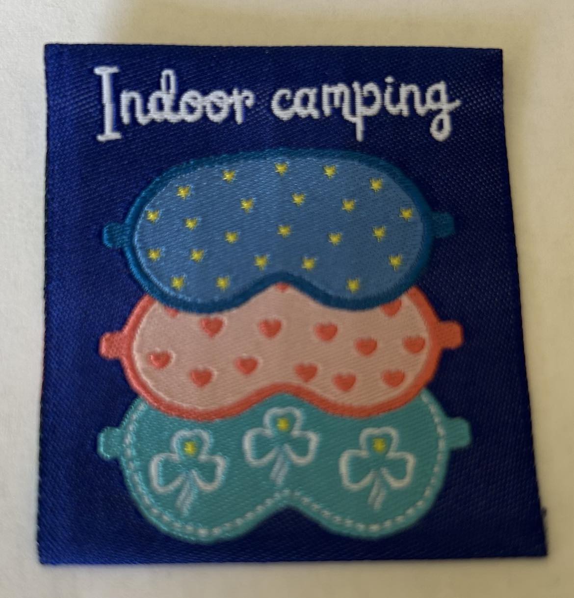 a square unbound badge with a blue background with the words indoor camping and three eye masks